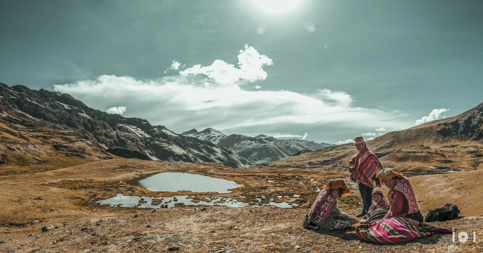 Peruvian family wearing traditional Andean clothing sitting on a mountain landscape with a lake and snow-capped mountains in the background.