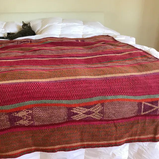Peruvian handwoven striped red wool blanket displayed on a bed with a cat next to it
